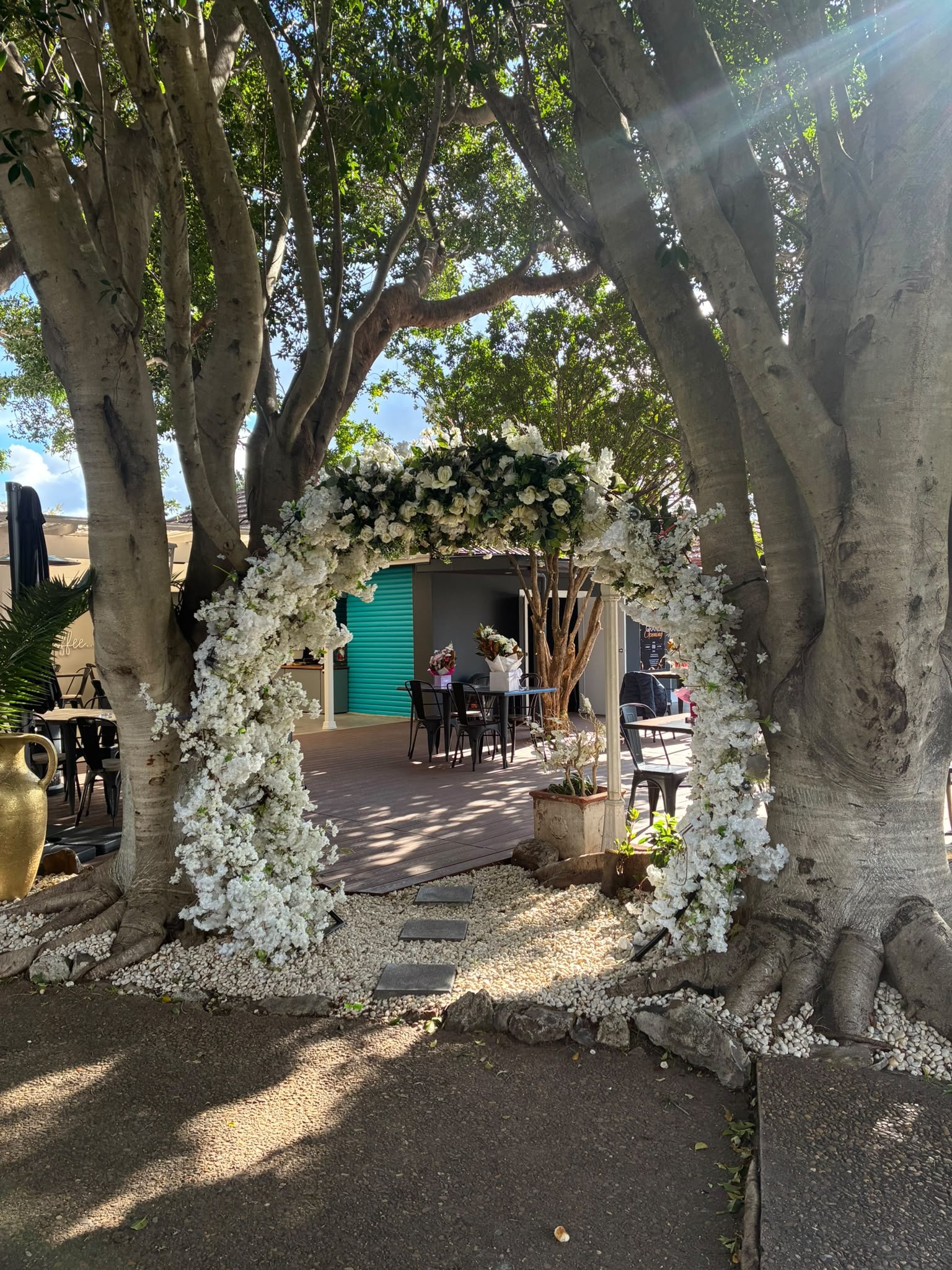 Breathtaking white floral archway nestled between majestic trees leading to outdoor garden dining