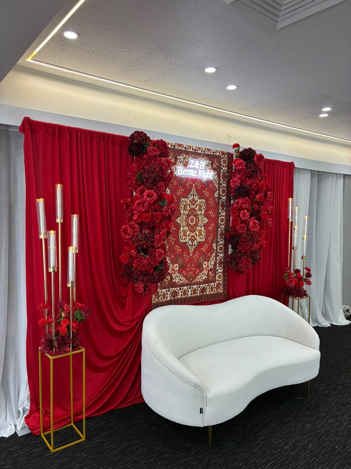 Luxurious Crimson Henna Night celebration stage with red velvet drapes, floral framing, curved sofa, and candlelight